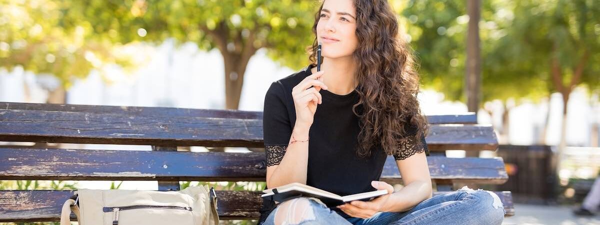 Cute Young Woman Sitting Outside In A Park Bench And Thinking While Writing In A Journal